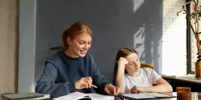 Two individuals sitting at a table with books and stationery, engaged in a study session. One person is pointing at a book while the other looks on.