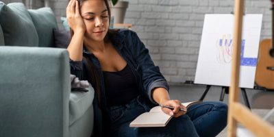 Person sitting on the floor next to a couch, holding a notebook and pen, with an easel and artwork in the background.