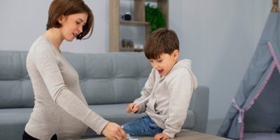 A pregnant woman and a young boy are playing a memory card game on a wooden table in a living room. The woman is standing and pointing at one of the cards, while the boy is sitting on the table, looking at the cards. The background includes a gray sofa, a bookshelf, and a play tent.