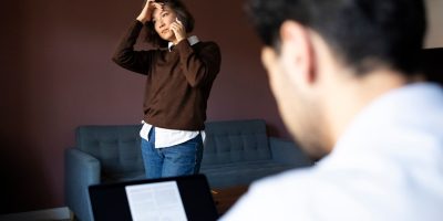 A person in a brown sweater and blue jeans stands in the background with hands on head, while another person in the foreground looks at a laptop screen displaying text.