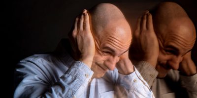 A person wearing a light-colored shirt is holding both hands to the sides of their head. The image has a motion blur effect, creating a double exposure appearance.