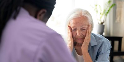 A person with long hair in a purple shirt is sitting across from an elderly person with white hair, who is holding their face with both hands. The elderly person appears to be in a state of distress. The background shows a bright room with a plant and some furniture.