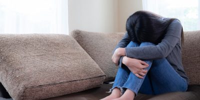 An emotional young woman sitting on a sofa, looking down with a pensive expression, conveying feelings of sadness and depression.