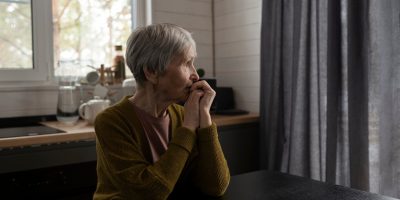 An elderly person with short gray hair sits at a dark table in a kitchen, wearing a mustard-colored cardigan over a pink shirt. The person is resting their chin on their hand, looking out of the window. The kitchen counter behind them has various kitchen items, including a coffee maker, cups, and a dish rack. A window with a view of trees and a curtain are also visible.