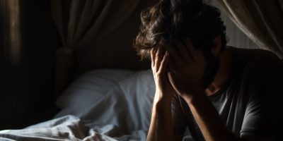 A person sitting on a bed with hands covering the face, appearing to be in distress. The room is dimly lit with curtains drawn and a bed with white sheets in the background.