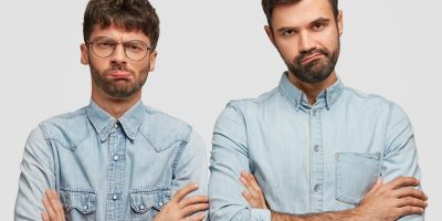 Two individuals standing side by side with their arms crossed, both wearing light blue denim shirts. Their faces are blurred.